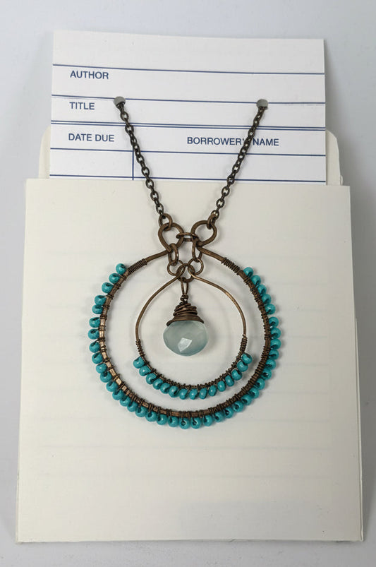 A vintage-style bronze pendant necklace with turquoise blue beads and a central round stone, displayed on an off-white card with a library or borrowing slip underneath.