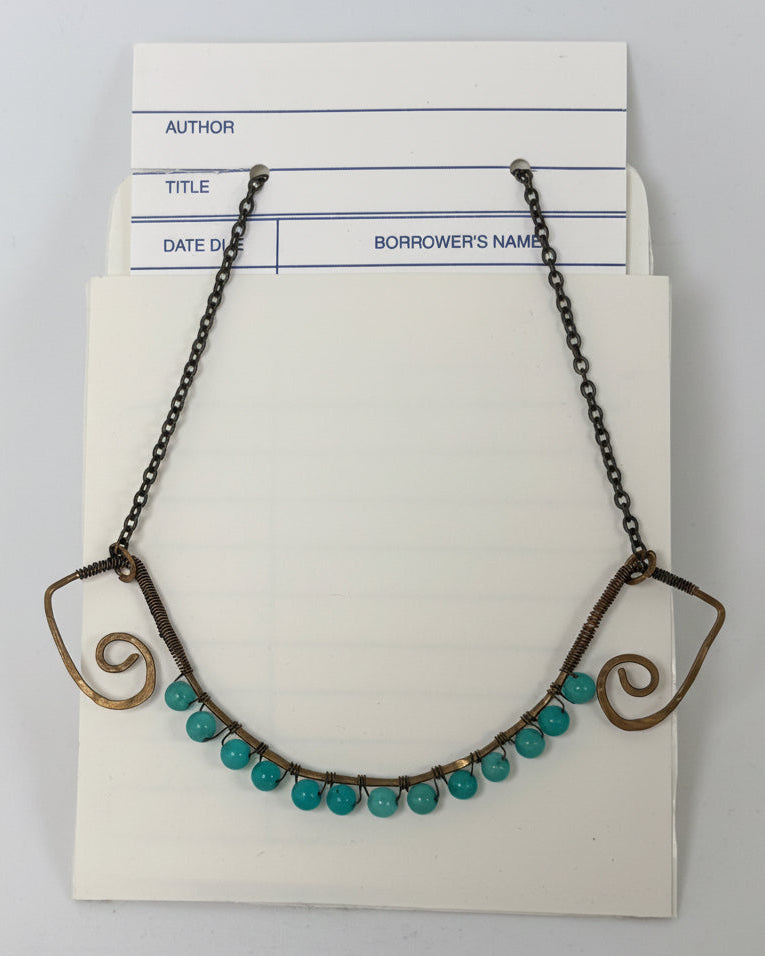 A vintage-style necklace with turquoise-colored round beads and dark metal scrollwork ends, displayed on a cream-colored card against a light gray background.