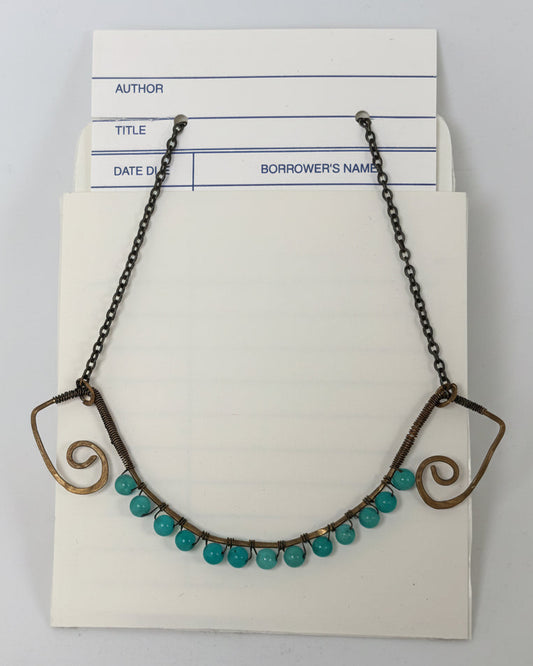 A vintage-style necklace with turquoise-colored round beads and dark metal scrollwork ends, displayed on a cream-colored card against a light gray background.
