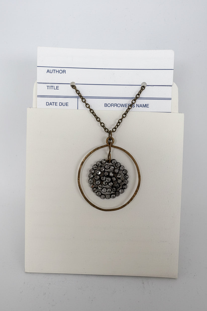 A bronze-finished necklace with a circular pendant featuring a dark gray beaded center, displayed on a cream-colored card with library loan forms tucked behind it.