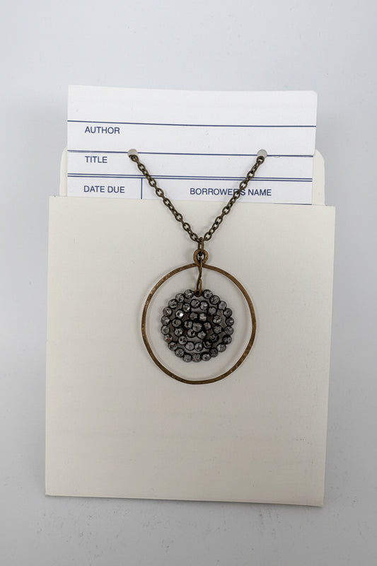 A bronze-finished necklace with a circular pendant featuring a dark gray beaded center, displayed on a cream-colored card with library loan forms tucked behind it.