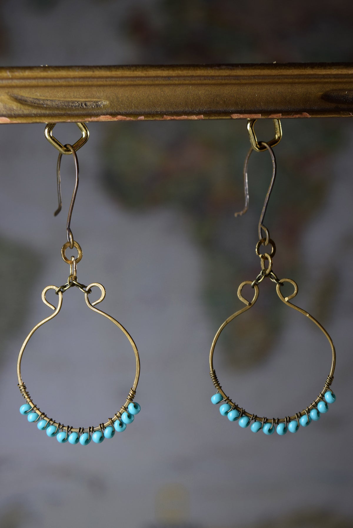 A pair of gold-toned drop earrings with small turquoise blue beads along the bottom curve, hanging from a distressed gold metal bar against a blurred gray background.