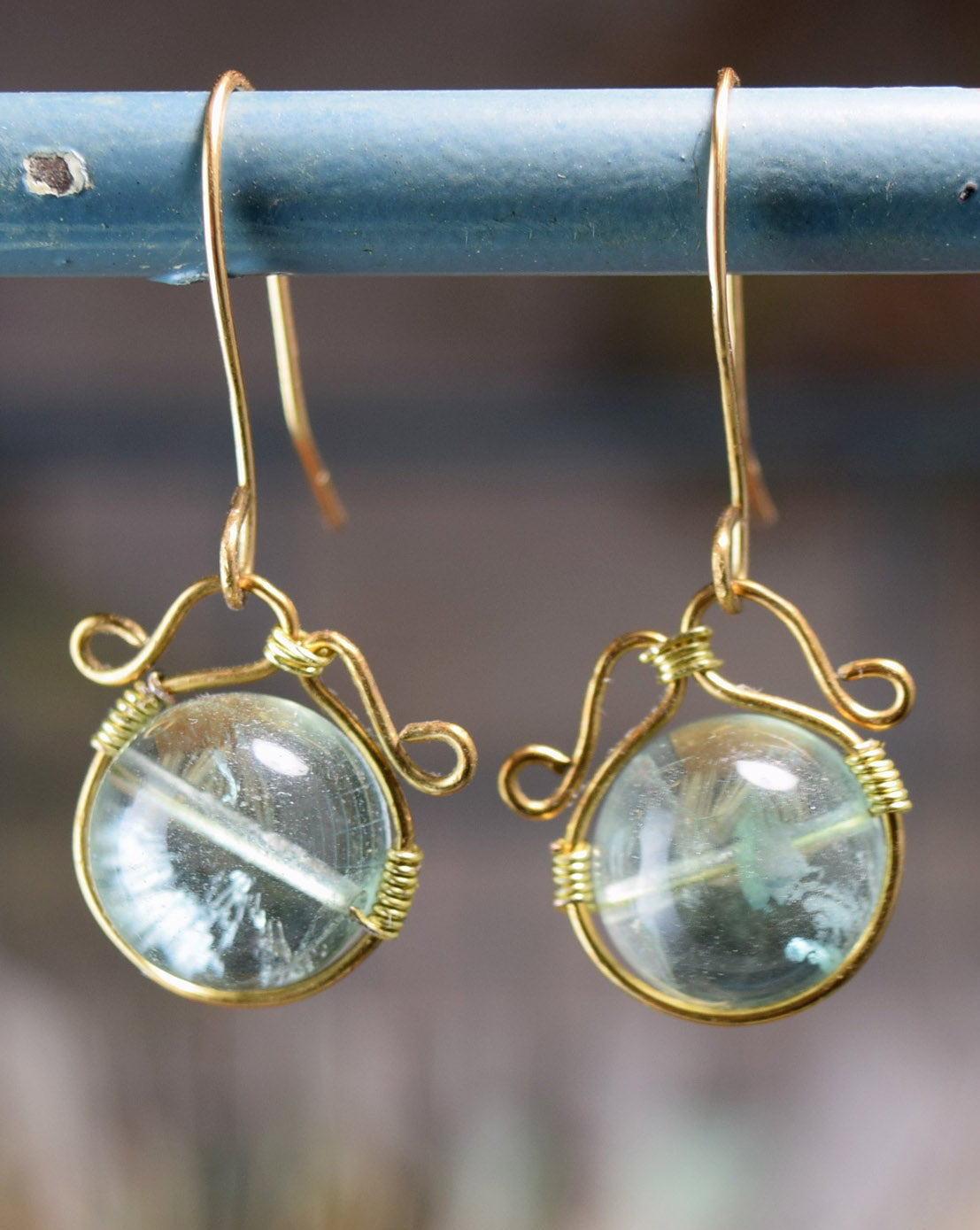 A pair of gold wire-wrapped dangle earrings with round clear gemstone pendants, hanging from a distressed blue metal fence.