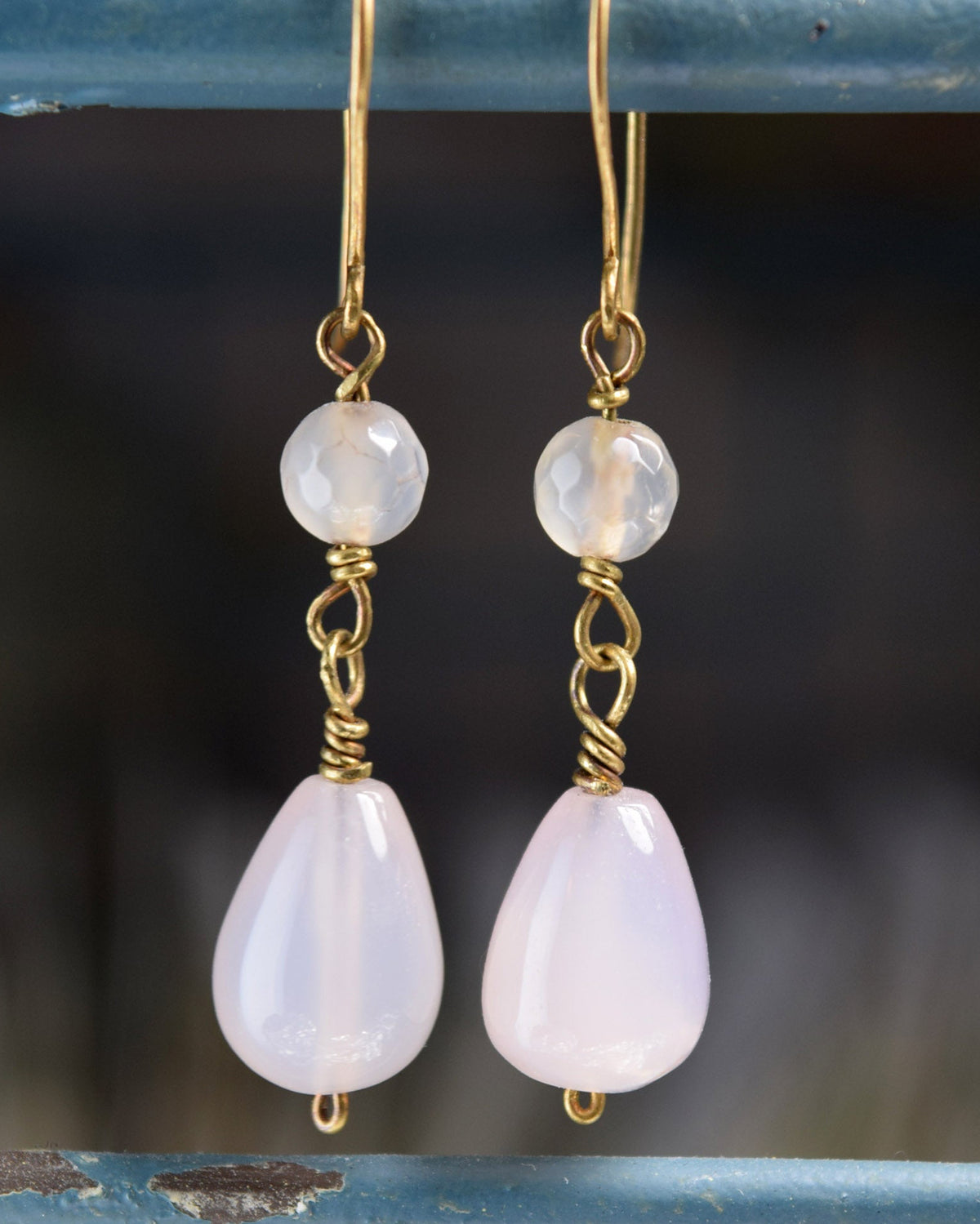 A pair of handmade dangle earrings featuring pale pink teardrop-shaped stones and round milky white beads, suspended from gold wire hooks and wrapped with gold metal, hanging against a rustic blue wooden background.
