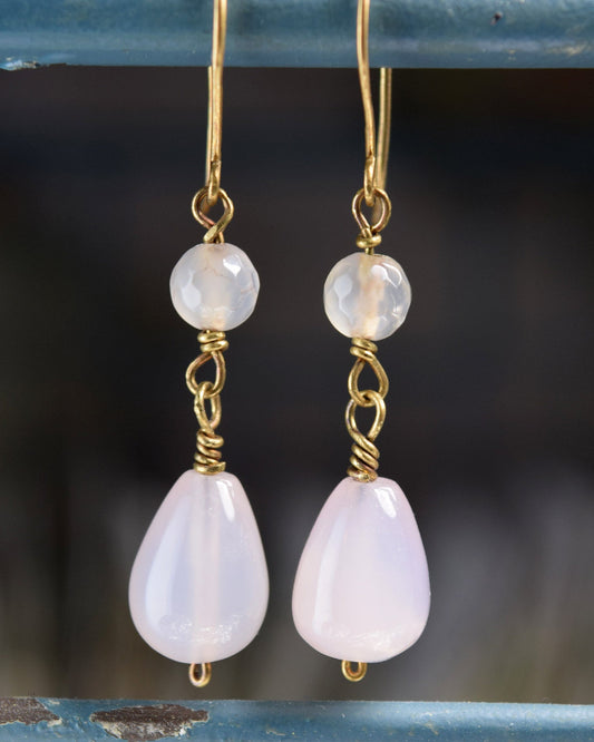 A pair of handmade dangle earrings featuring pale pink teardrop-shaped stones and round milky white beads, suspended from gold wire hooks and wrapped with gold metal, hanging against a rustic blue wooden background.