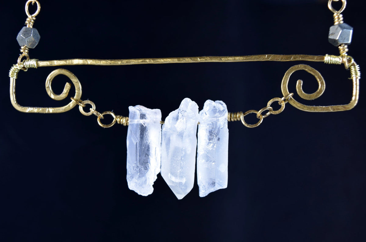 A gold-toned metal necklace featuring three raw, clear quartz crystal points suspended from a horizontal bar, set against a solid black background.
