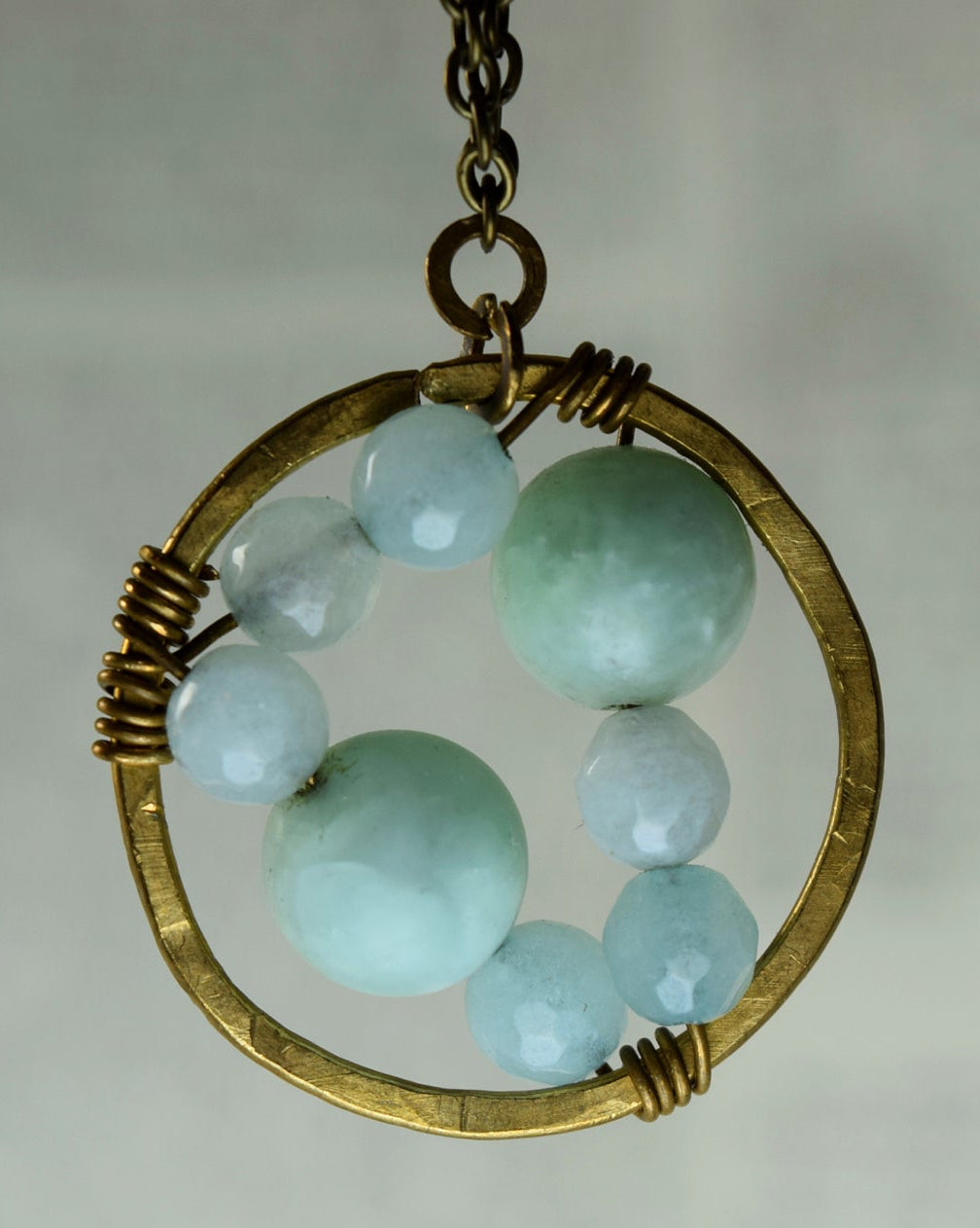 A handmade pendant necklace featuring light blue faceted beads and a gold-toned circular frame with wire wrapping, hanging against a soft, blurred light gray background.